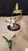 (2) MARURI HUMMINGBIRD FINE  PORCELAIN AND BRONZE STUDIO DESIGN H8905 ANNA'S HUMMINGBIRD WITH LILY, CIRCA 1989, BROKEN LIMB AND A RUBY THROATED HUMMINGBIRD WITH ORCHAD, LEAF MISSING OR BIRD, WING BROKEN - (2) HUMMINGBIRDS - (GREAT ROOM)